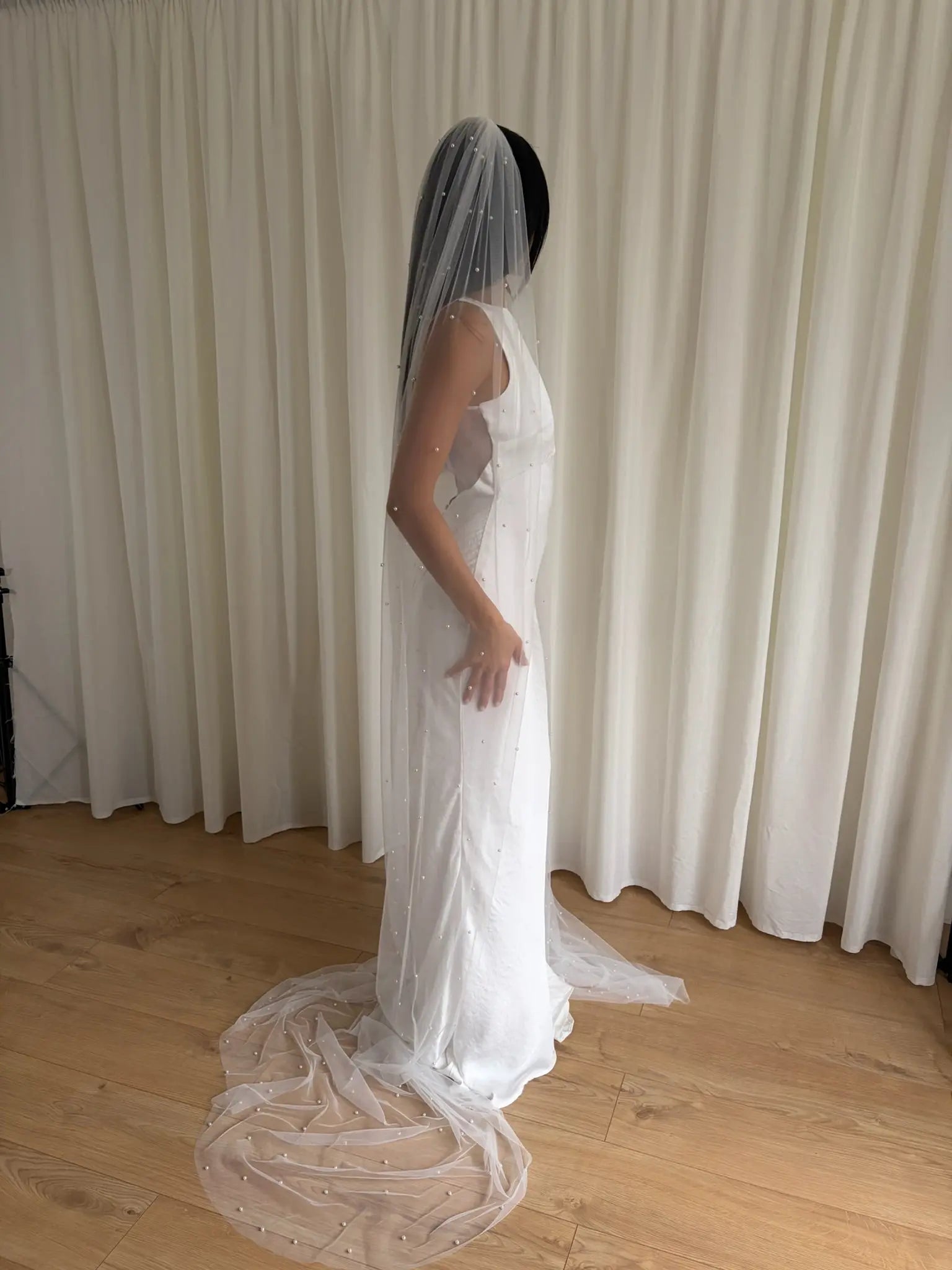 Person wearing a white wedding dress with a long pearl veil against a plain background