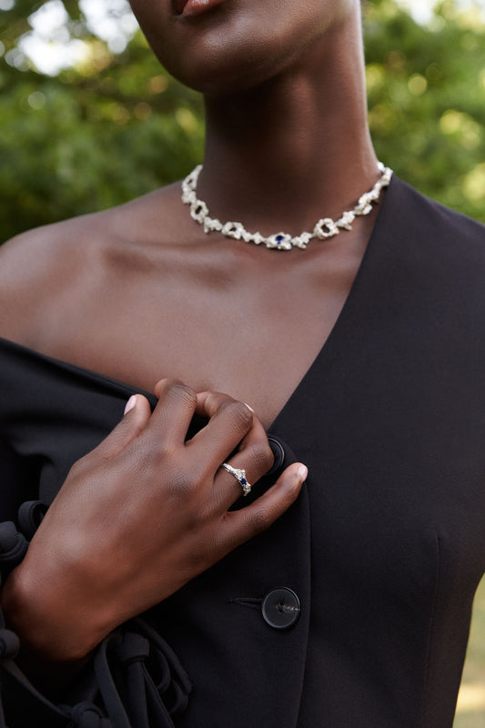 Person wearing a black outfit with a necklace and ring, standing against a blurred green background