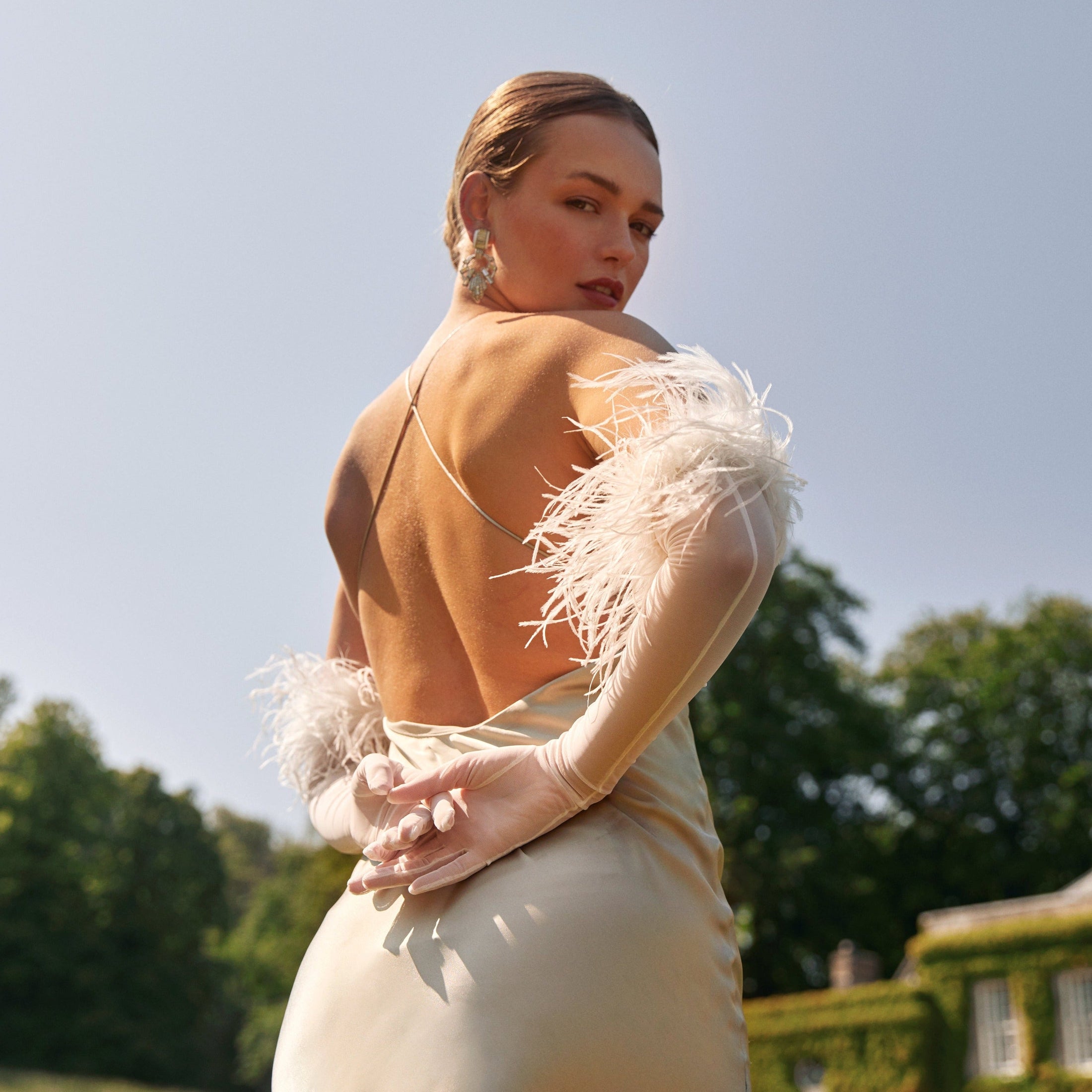 Woman in a beige dress with feather bridal gloves standing outdoors against a clear sky.