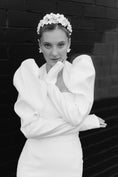 Load image into Gallery viewer, Woman in a white dress with puffed sleeves and floral headpiece against a dark brick wall.
