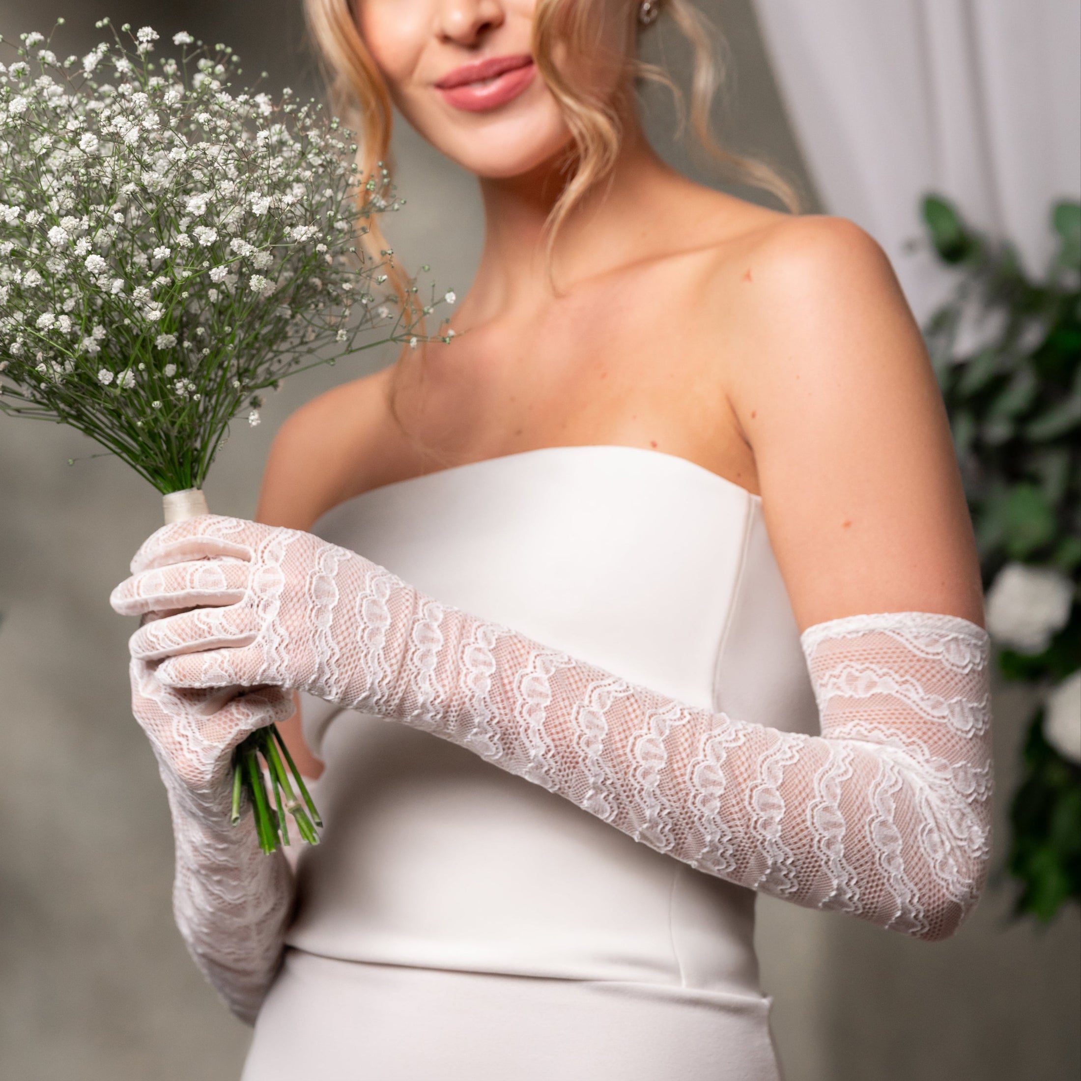 Bride wearing a white dress and lace gloves holding a bouquet of small white flowers.