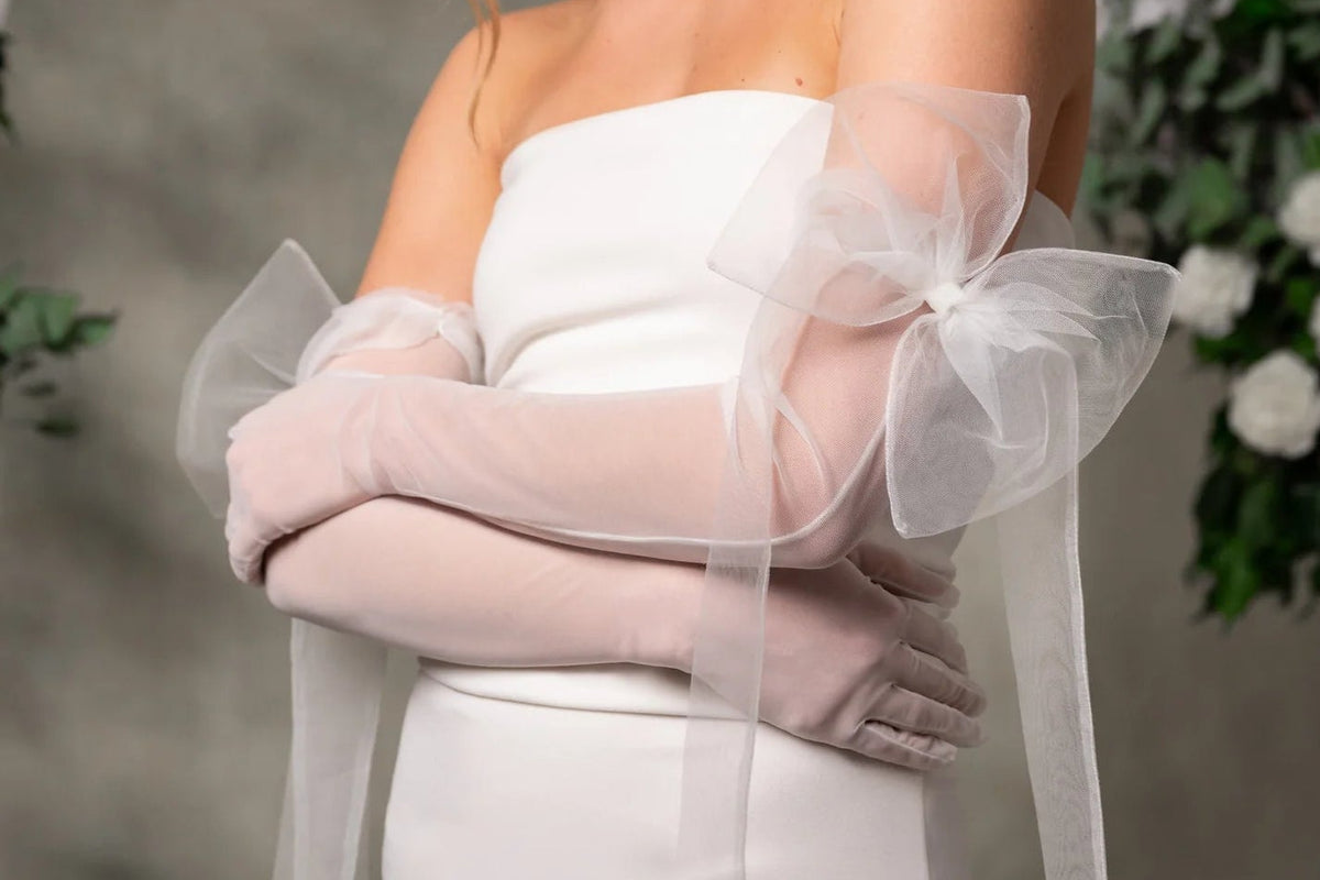 A woman wearing a strapless white dress and long sheer blush pink gloves with oversized bow details on the sleeves, posed indoors against a soft gray backdrop with floral decor.