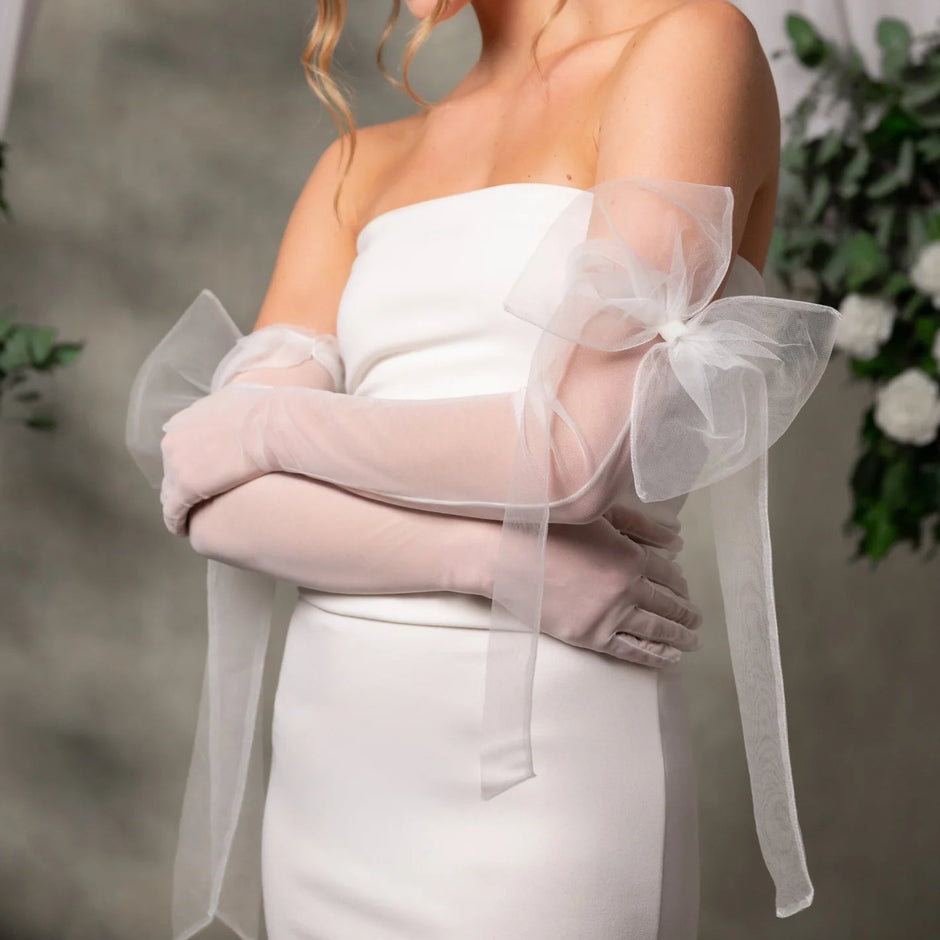 A woman wearing a strapless white dress and long sheer blush pink gloves with oversized bow details on the sleeves, posed indoors against a soft gray backdrop with floral decor.