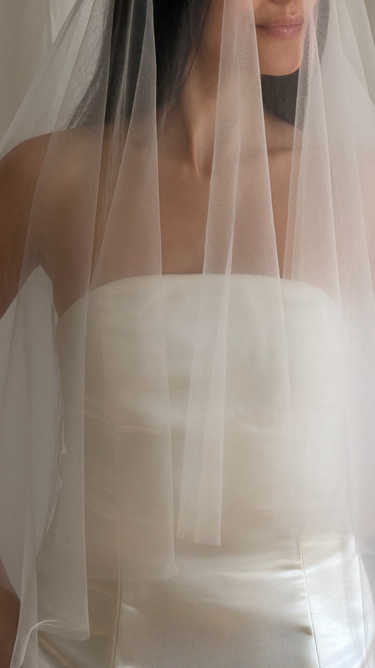 Bride wearing a white wedding dress with a plain veil against a white curtain background.