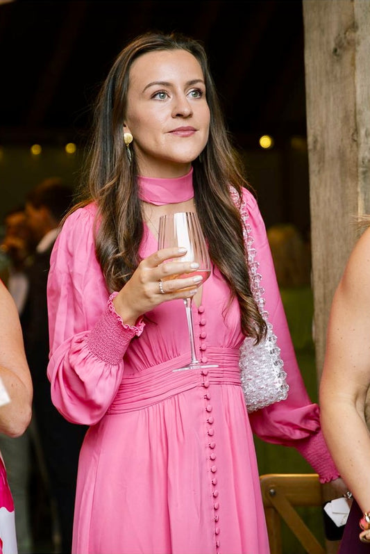 Woman in a pink dress holding a glass of wine at an outdoor event wearing a bridal bag