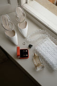 Load image into Gallery viewer, White high-heeled shoes, a ring, earrings, a perfume bottle, and a bridal bag on a light surface near a window.

