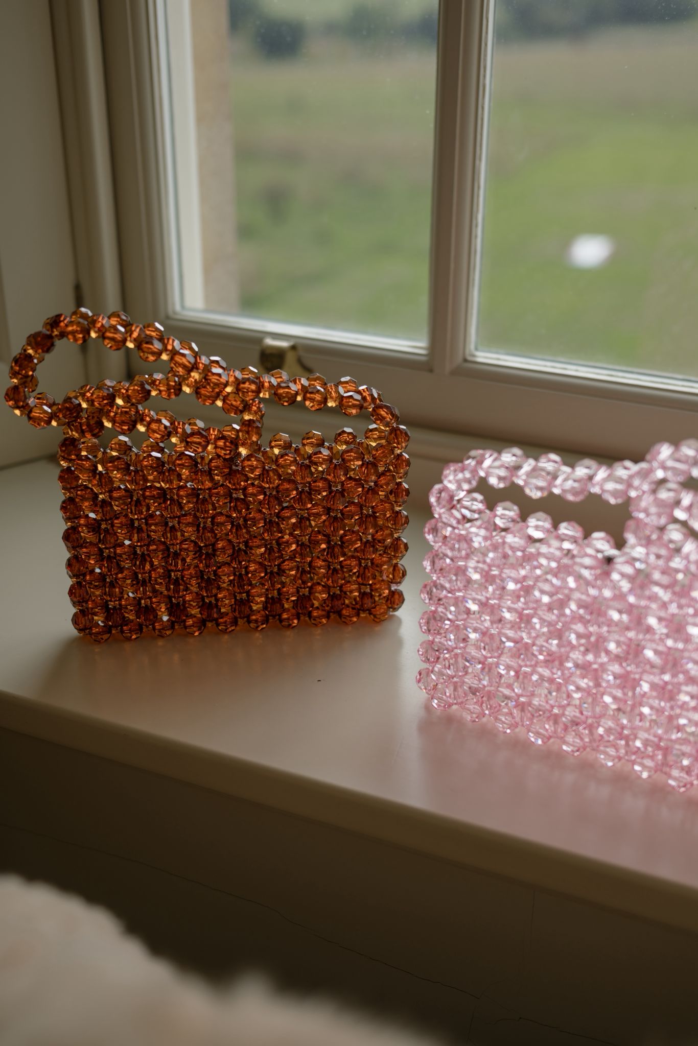 Two beaded bridal bags, one orange and one pink, on a windowsill with a view of greenery outside.