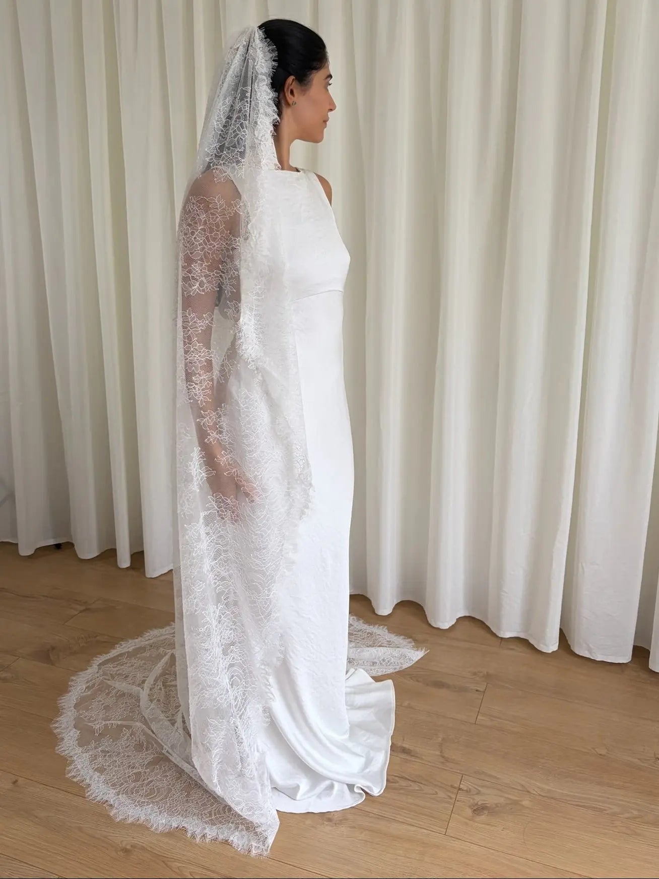 Woman wearing a white lace veil and wedding dress against a white curtain background