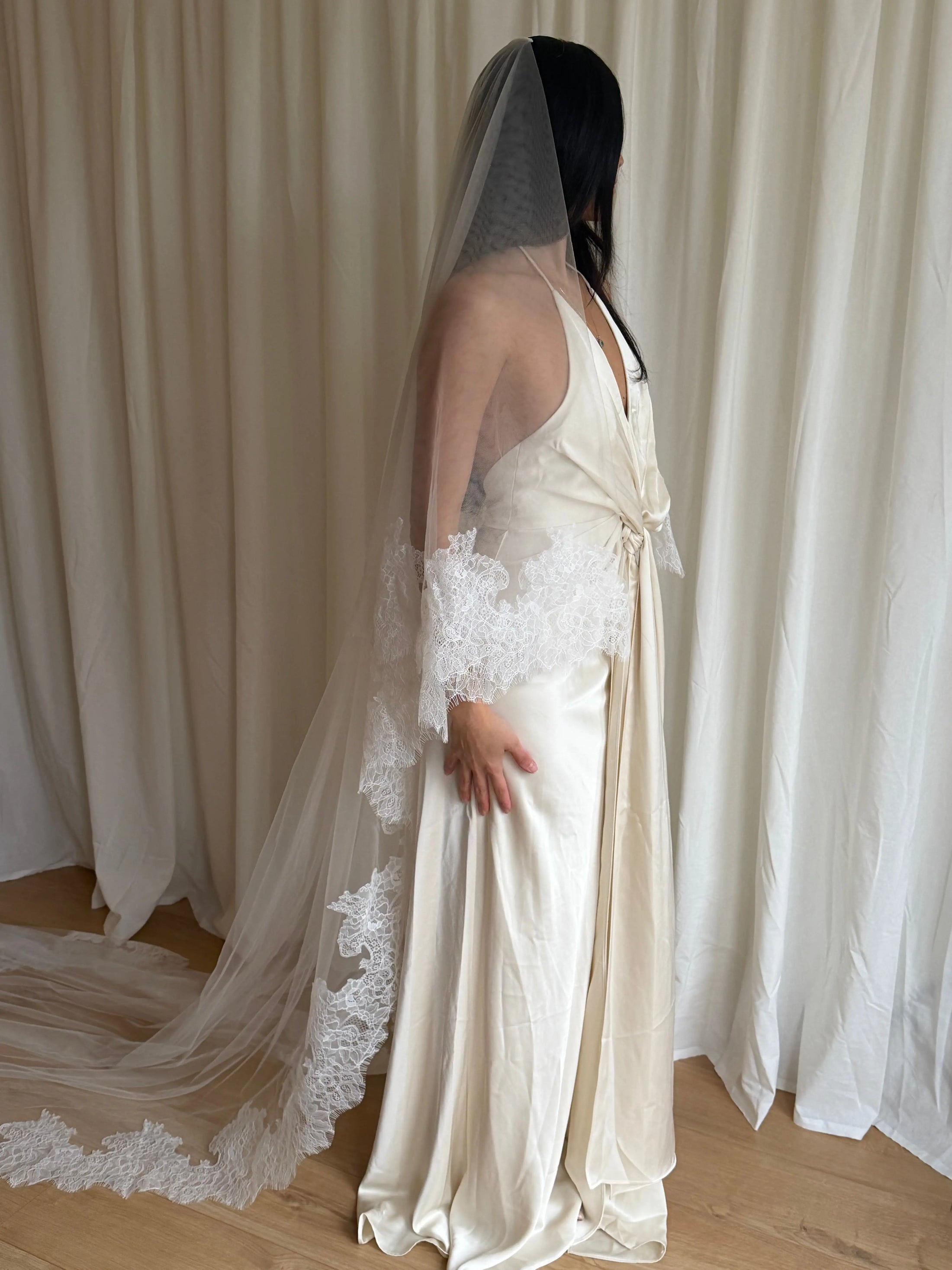 Bride wearing a white wedding dress with a lace veil against a white curtain background.