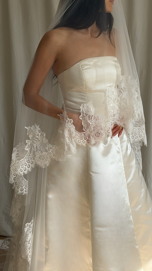 Bride wearing a white wedding dress with a lace veil against a white curtain background.