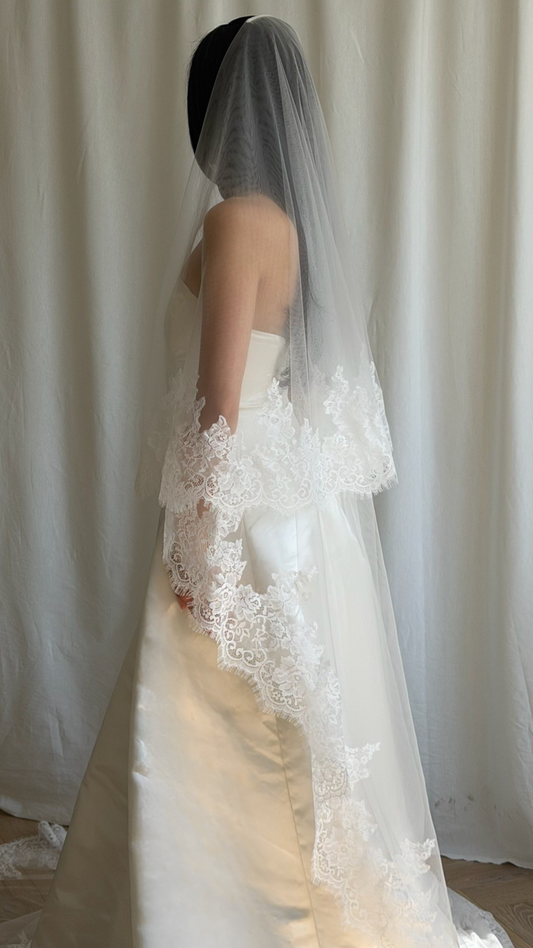 Bride wearing a white wedding dress with a lace veil against a white curtain background.