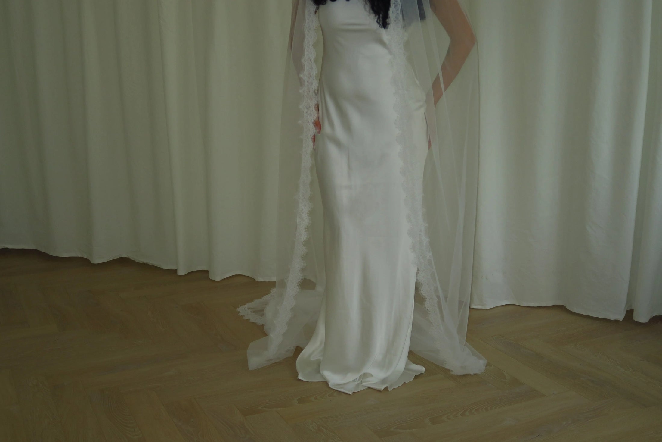 Person wearing a white wedding dress with a long veil in front of white curtains.