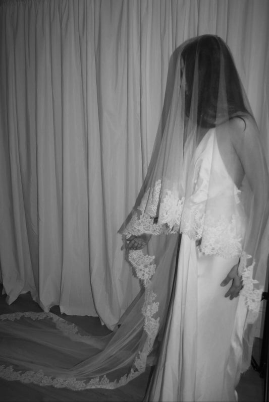 Black and white photo of a person in a wedding dress with a long lace trim veil against a curtain backdrop.