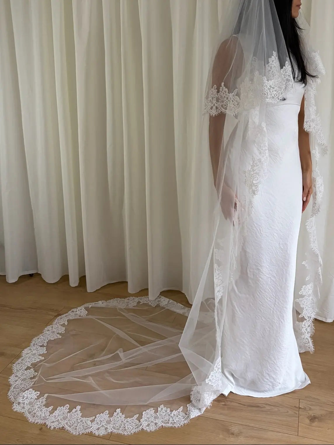White wedding dress with a long lace veil in front of a white curtain.