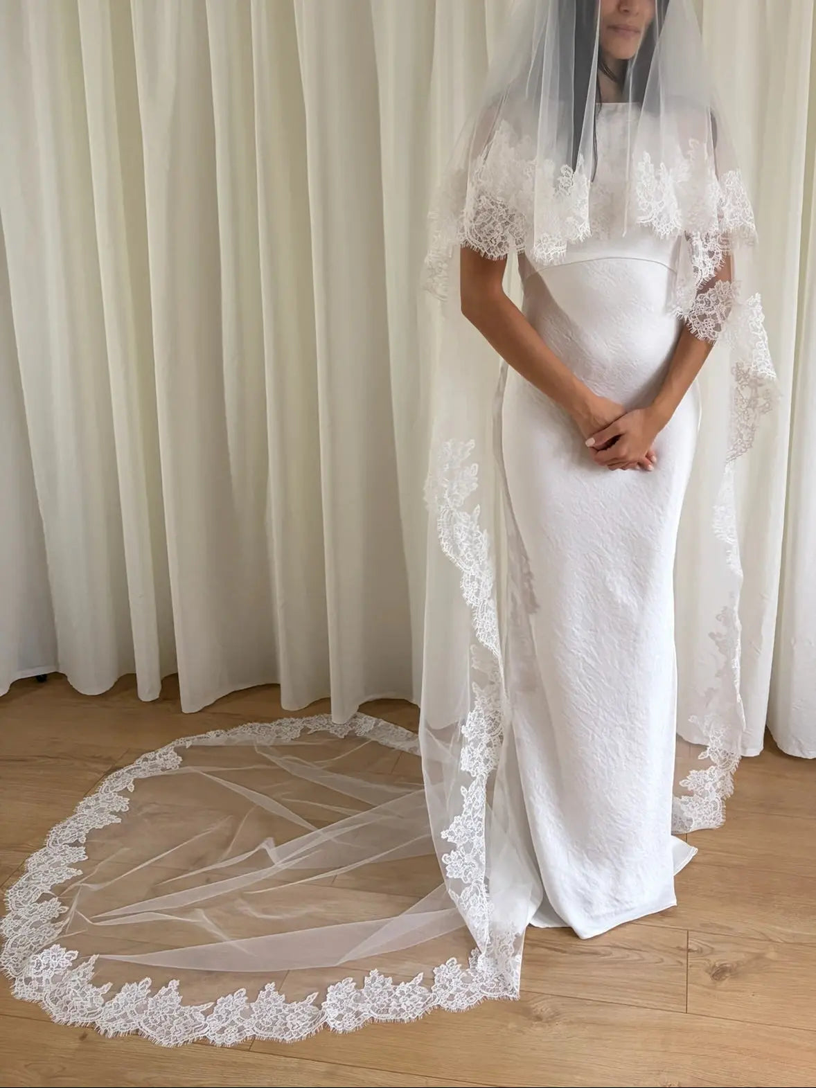 White wedding dress with a long lace veil in front of a white curtain.