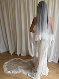 Load image into Gallery viewer, A woman shown from behind wearing a long white lace-trimmed tulle bridal veil over a cream wedding dress, standing on a wooden floor in front of draped cream fabric.
