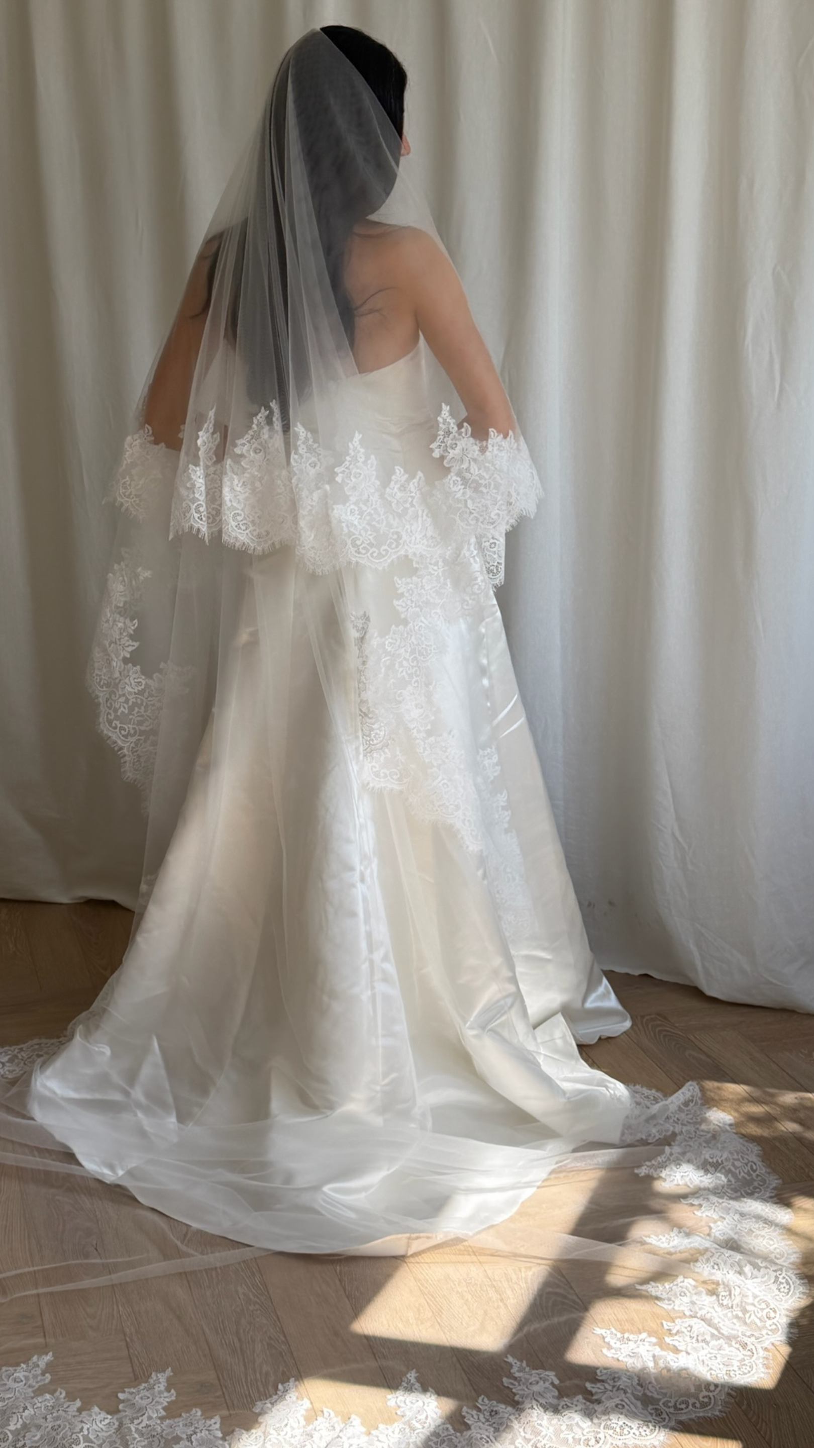 Bride wearing a white wedding dress with a lace veil against a white curtain background.