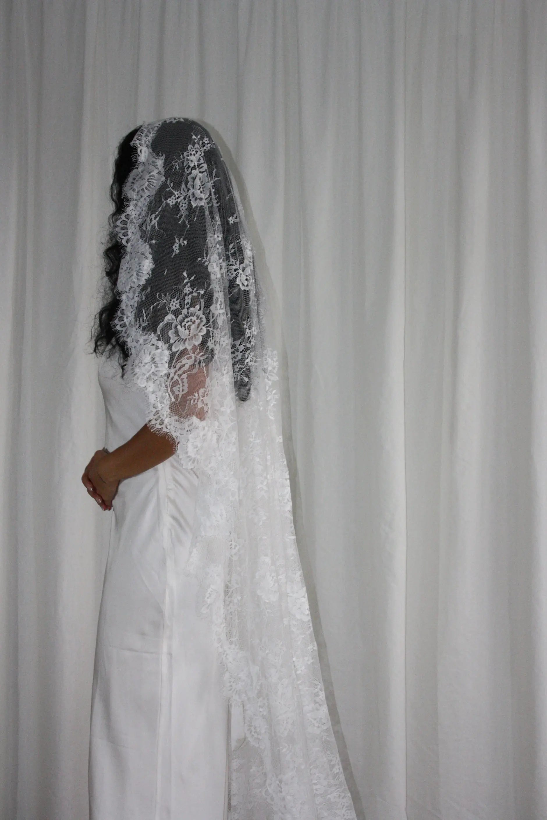 Person wearing a white wedding dress with a long lace veil against a plain background