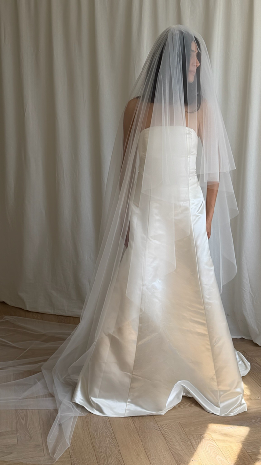 Bride in a white wedding dress with a long veil standing against a white curtain.