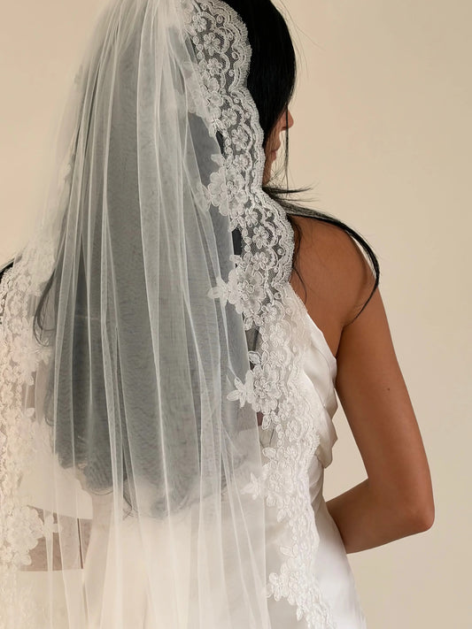 A bridal veil with intricate white lace trim worn by a model, shown from the back against a plain beige background.