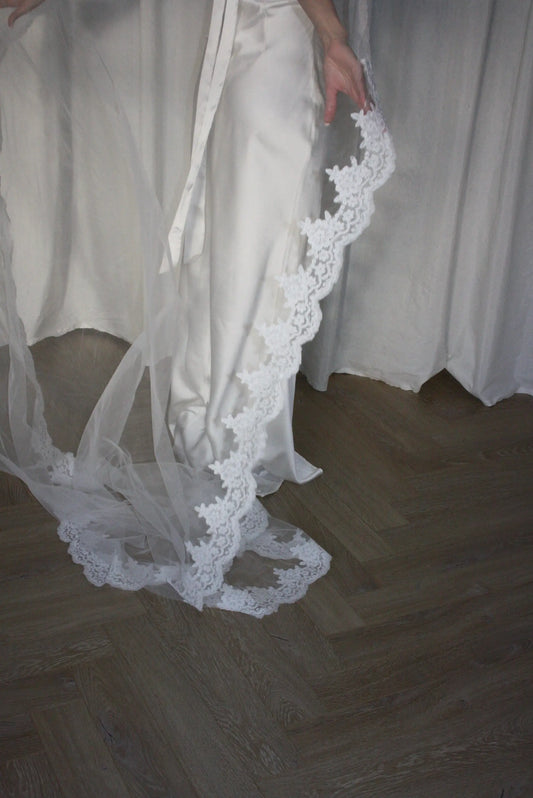 A white satin bridal gown with a long sheer veil featuring a scalloped white lace border, shown on a model standing on a wooden herringbone floor.