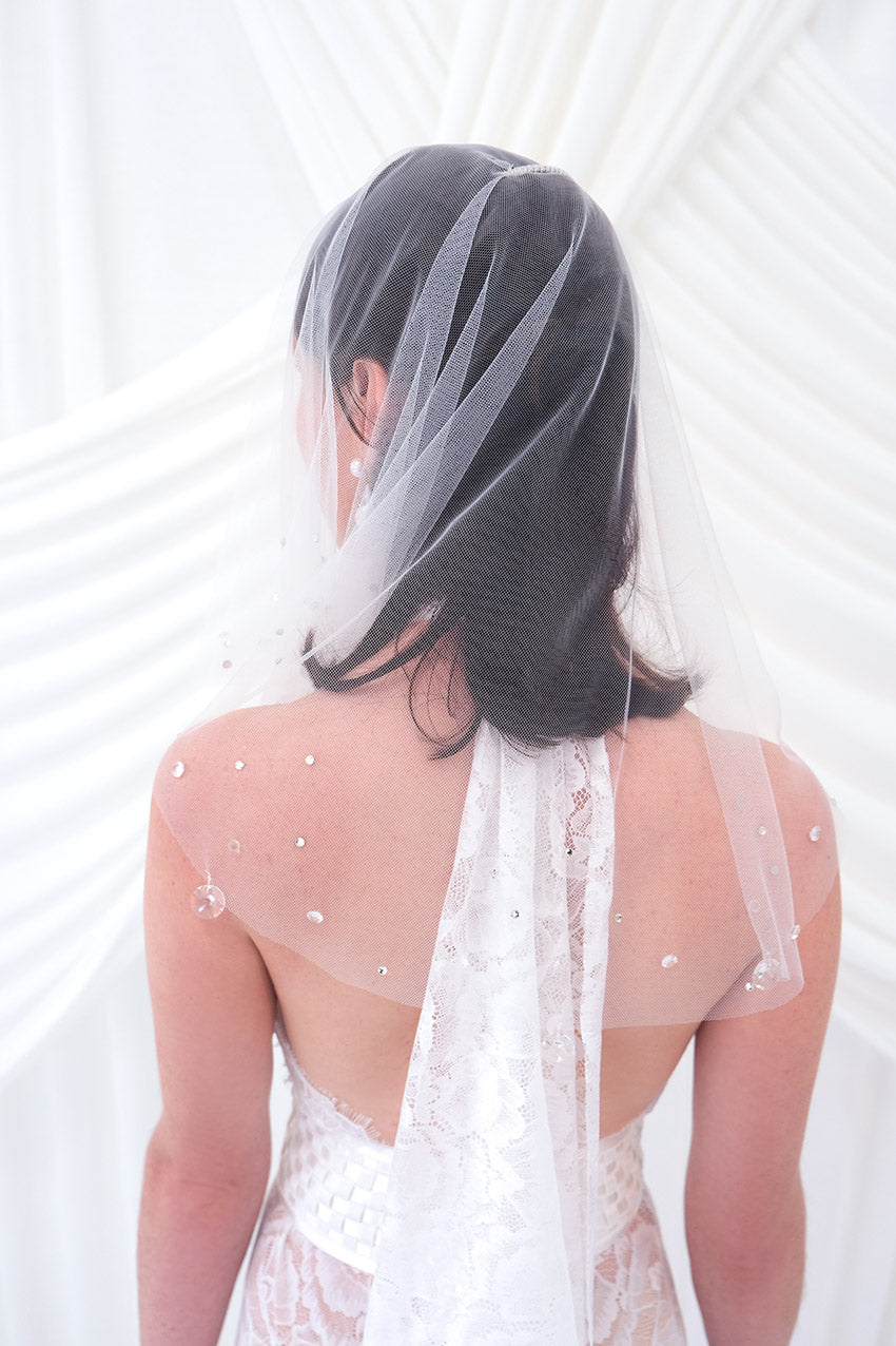 A bride shown from the back wearing a sheer white tulle bridal veil with delicate bead and sequin embellishments, paired with a white wedding dress against a soft white draped backdrop.