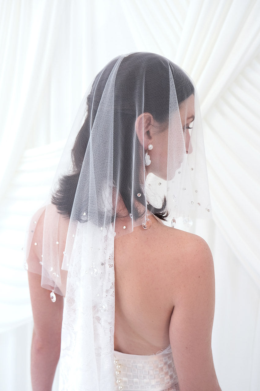 A bride shown from the back and side wearing a sheer white tulle bridal veil with delicate bead and sequin embellishments, paired with a white wedding dress against a soft white draped backdrop.