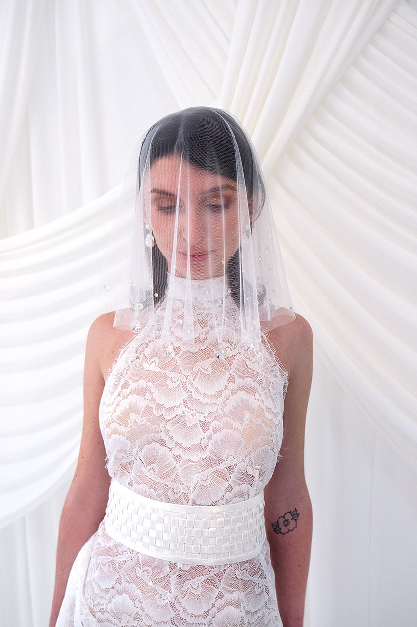 A bride shown from the front wearing a sheer white tulle bridal veil with delicate bead and sequin embellishments, paired with a white wedding dress against a soft white draped backdrop.