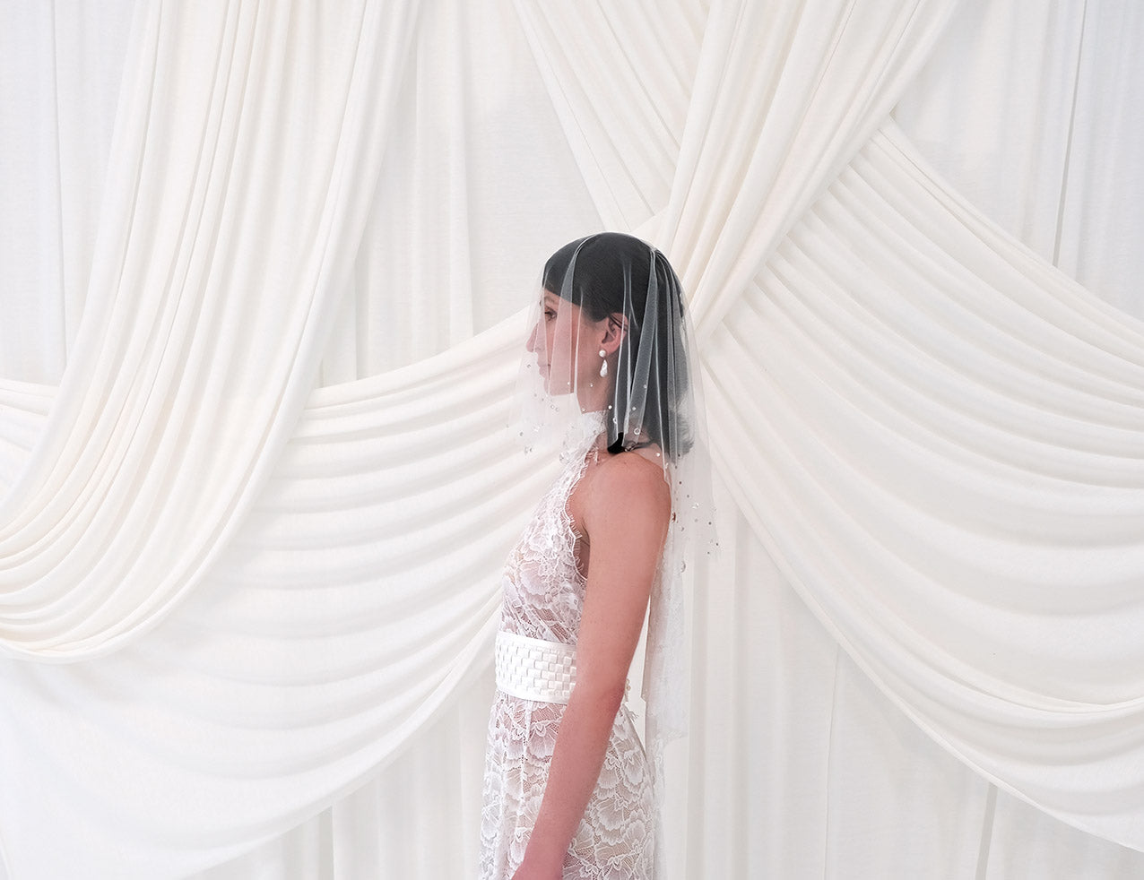 A woman in profile wearing a sheer crystal bridal veil and a white lace wedding dress, standing in front of draped white fabric.