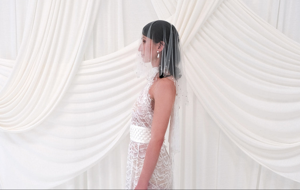 A woman in profile wearing a sheer crystal bridal veil and a white lace wedding dress, standing in front of draped white fabric.