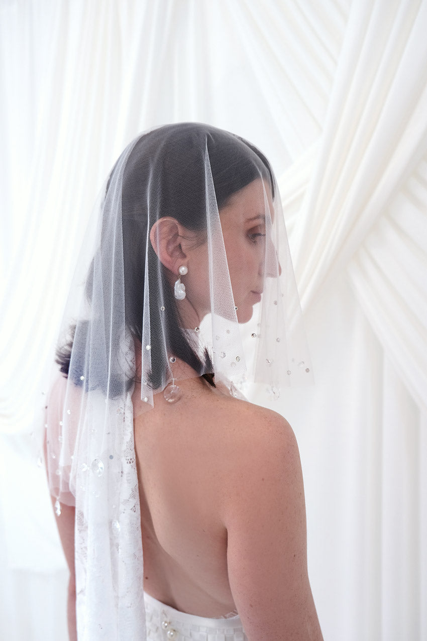 A bride shown from the back and side wearing a sheer white tulle bridal veil with delicate bead and sequin embellishments, paired with a white wedding dress against a soft white draped backdrop.