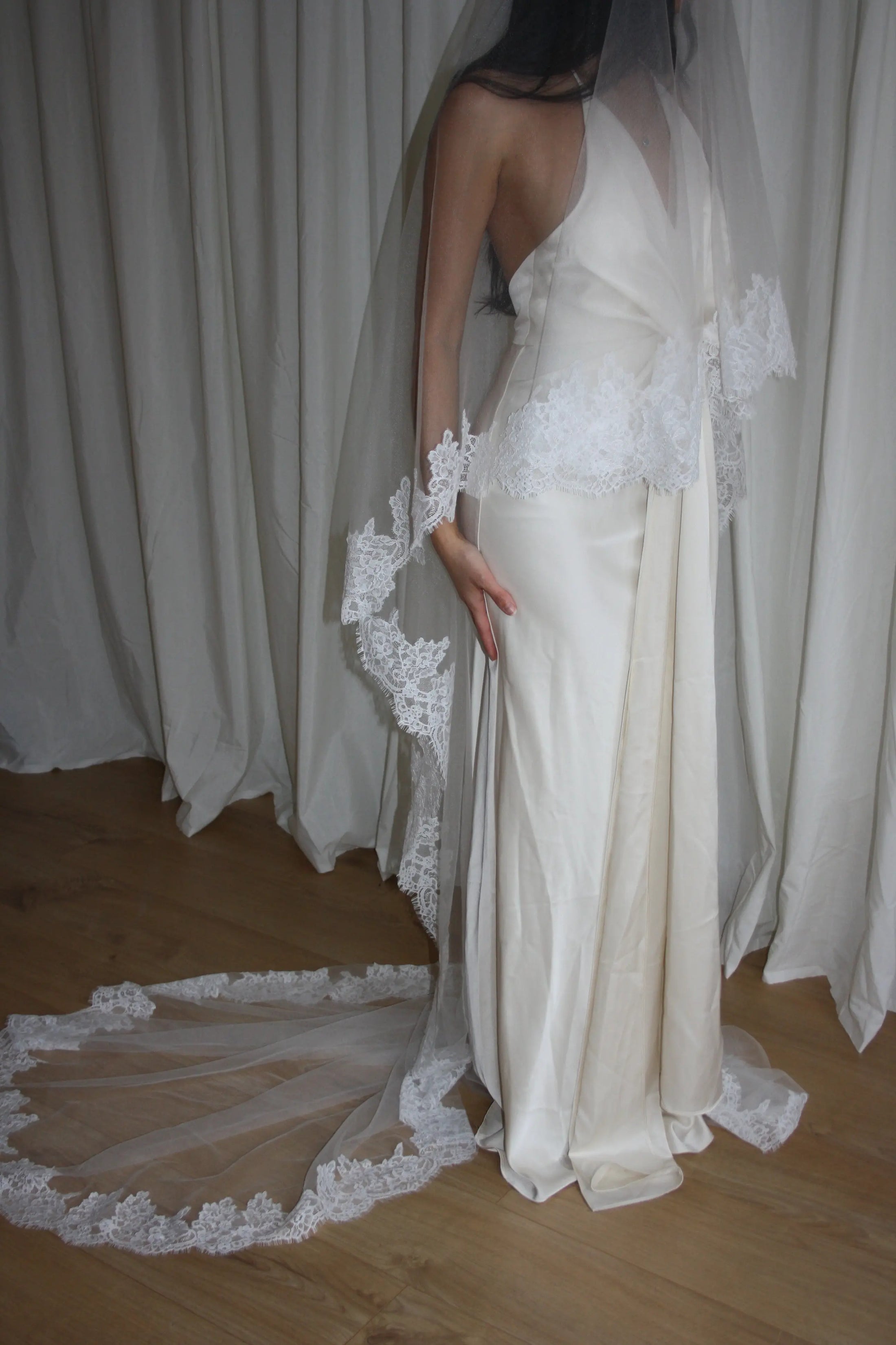 person in a wedding dress with a long lace trim veil against a curtain backdrop.
