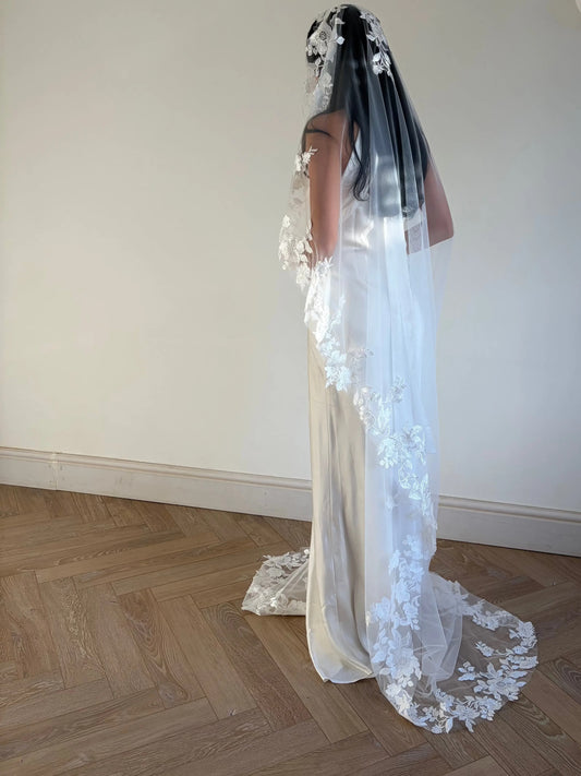 A woman wearing a long white bridal veil with floral lace appliqué and a matching sleeveless wedding dress, standing in a room with a light beige wall and wood herringbone floor.