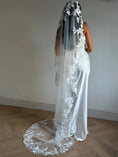Load image into Gallery viewer, A woman wearing a long white bridal veil with floral lace applique, shown from the back in a room with a light gray wall and wood herringbone floor.
