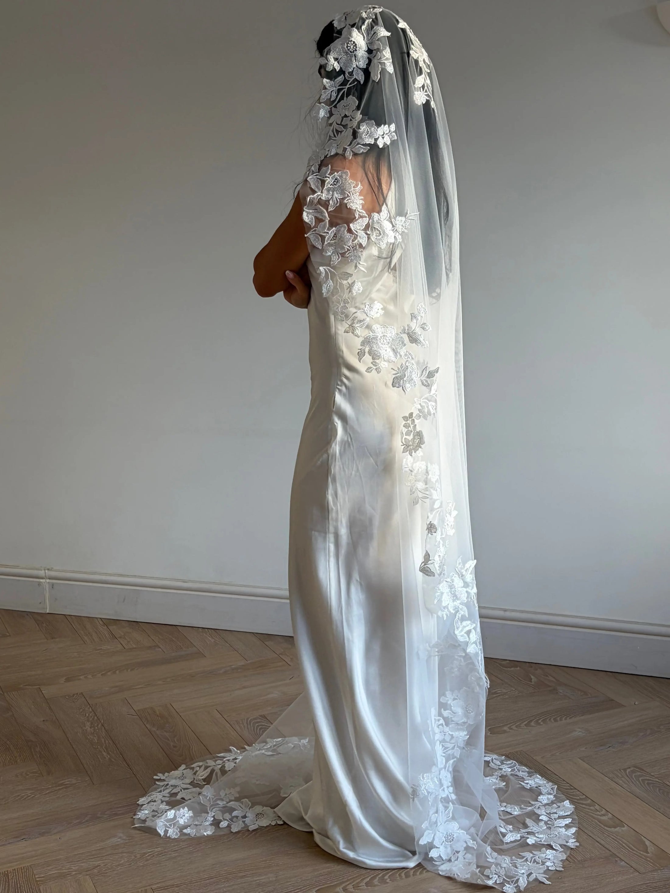 A woman wearing a long white bridal veil with floral lace appliqué and a matching sleeveless wedding dress, standing in a room with a light beige wall and wood herringbone floor.
