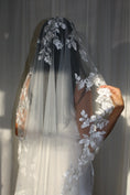 Load image into Gallery viewer, A woman wearing a long white bridal veil with floral lace appliqué and a matching sleeveless wedding dress, with a white curtain background.
