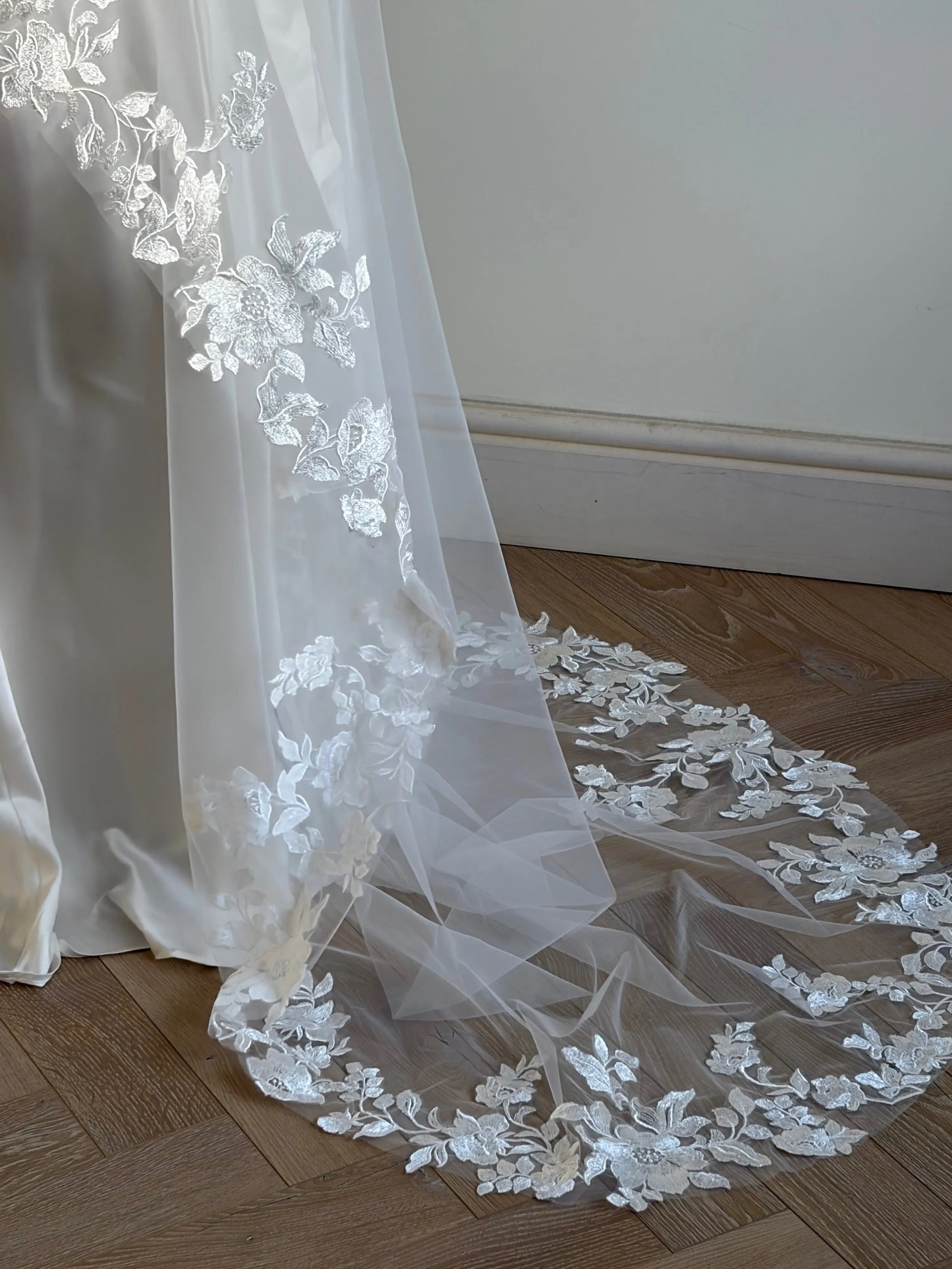 White floral lace bridal veil draped over a white satin dress, spread out on a wooden parquet floor against a neutral wall.