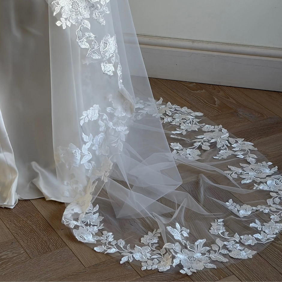 White floral lace bridal veil draped over a white satin dress, spread out on a wooden parquet floor against a neutral wall.