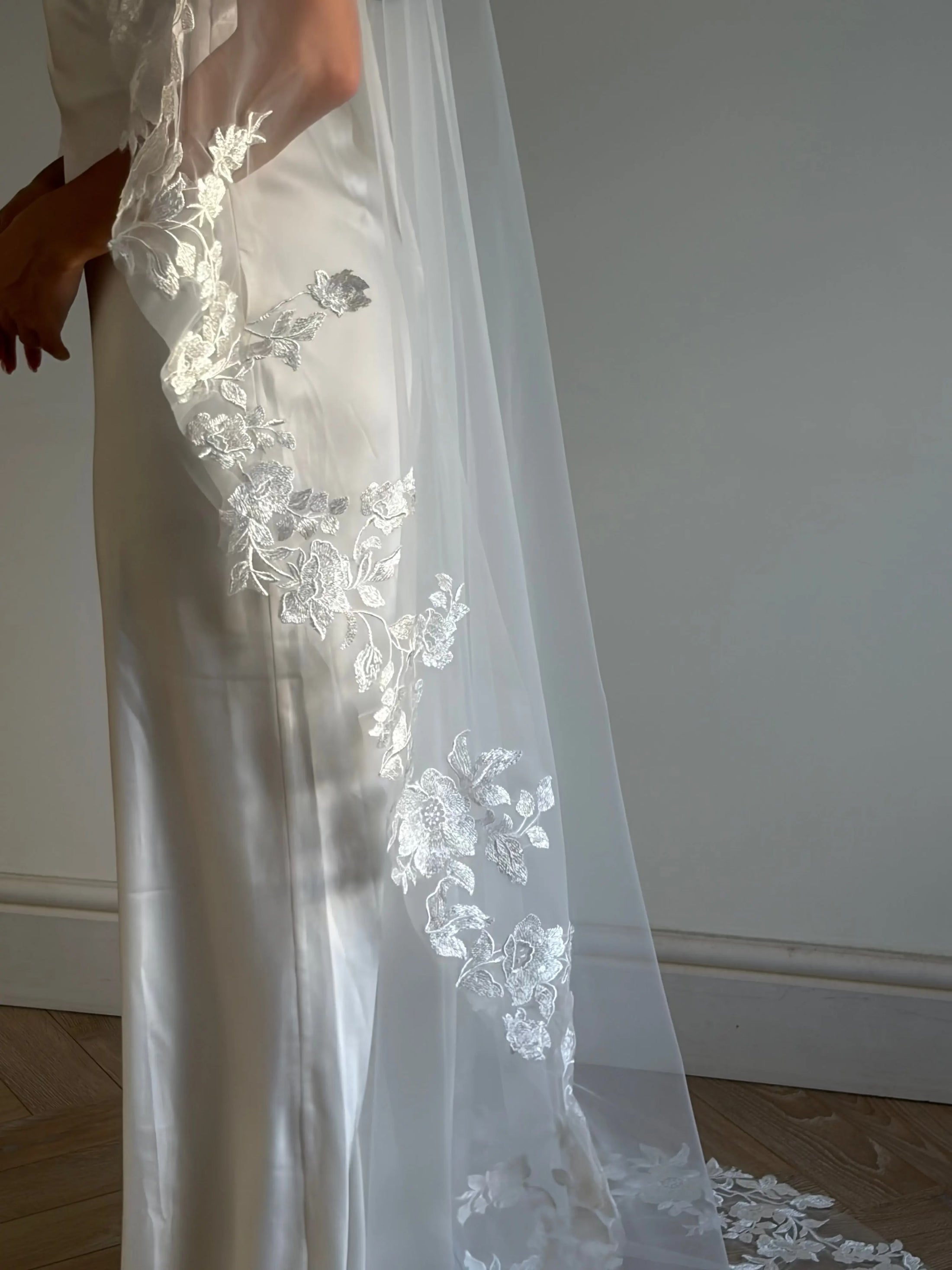 A close-up of a white satin wedding dress with a long sheer tulle veil featuring intricate white floral lace applique, worn by a model standing indoors against a neutral gray wall.