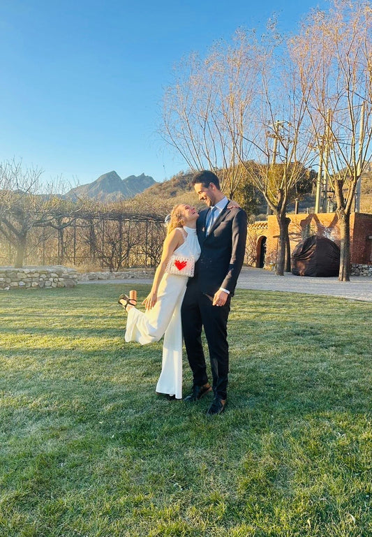 Man and woman in formal attire standing on grass with mountains in the background wearing bridal bag