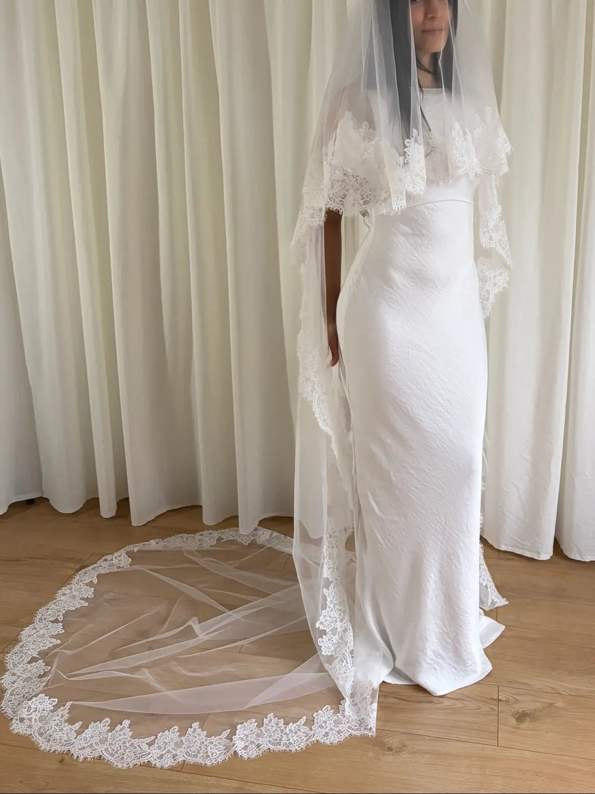 White wedding dress with a long lace veil in front of a white curtain.