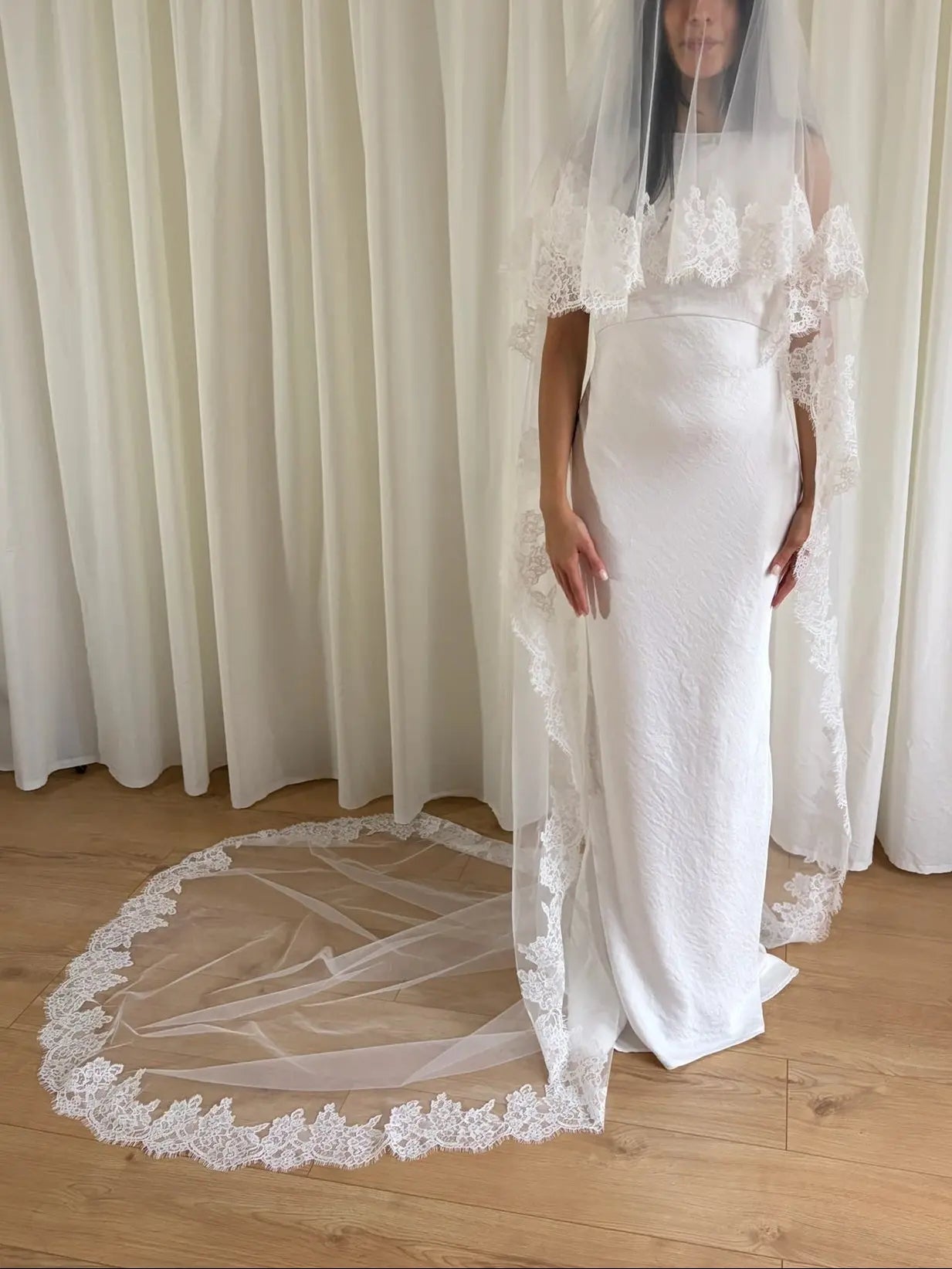 White wedding dress with a long lace veil in front of a white curtain.