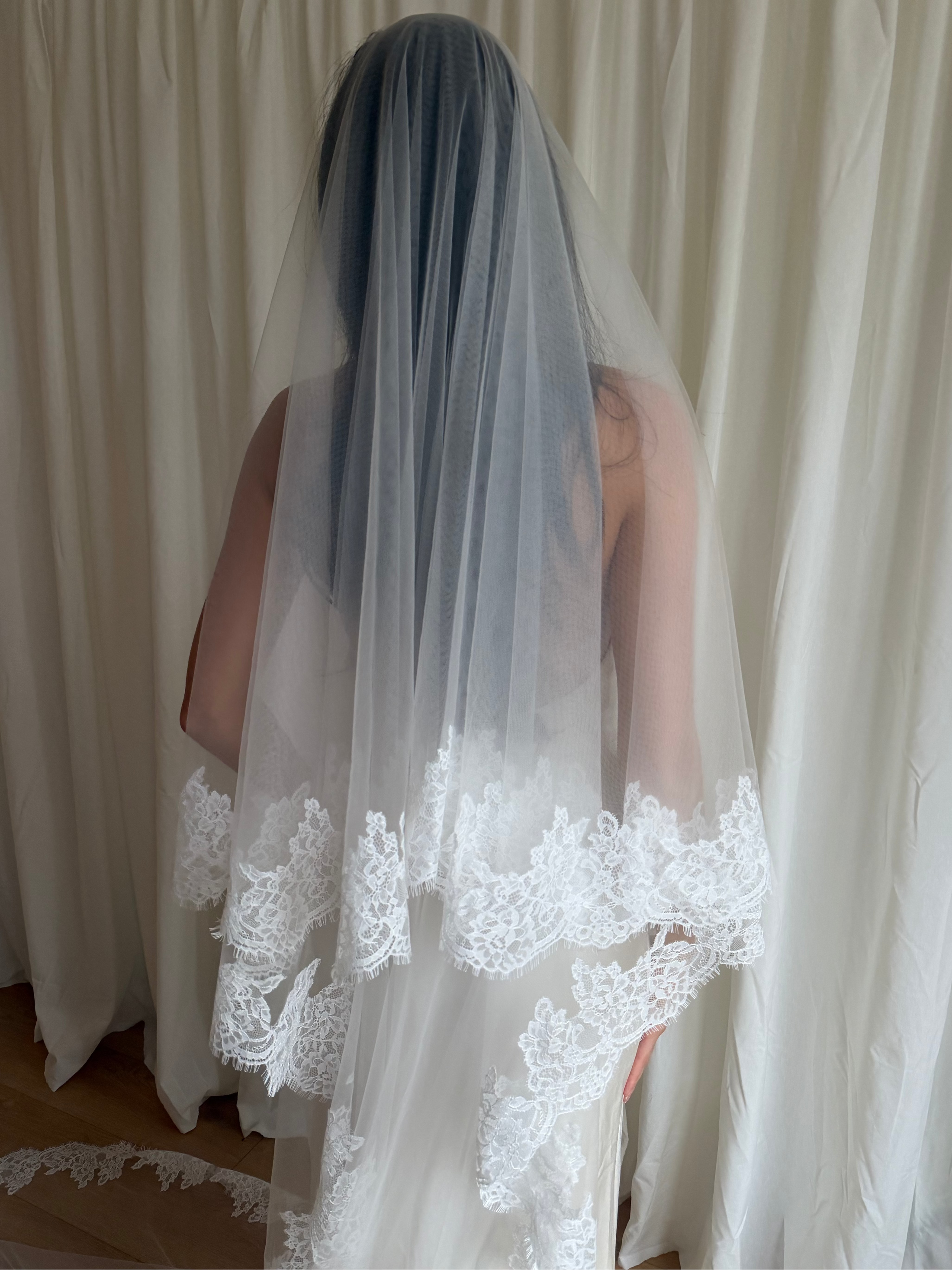 A bride viewed from behind wearing a long, sheer white tulle bridal veil with intricate white lace trim along the bottom, standing in front of cream-colored curtains.