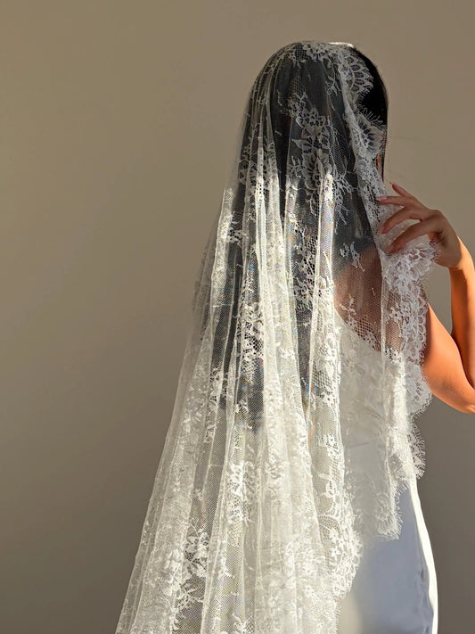 A long white lace bridal veil worn by a model, shown from the back against a plain beige wall.