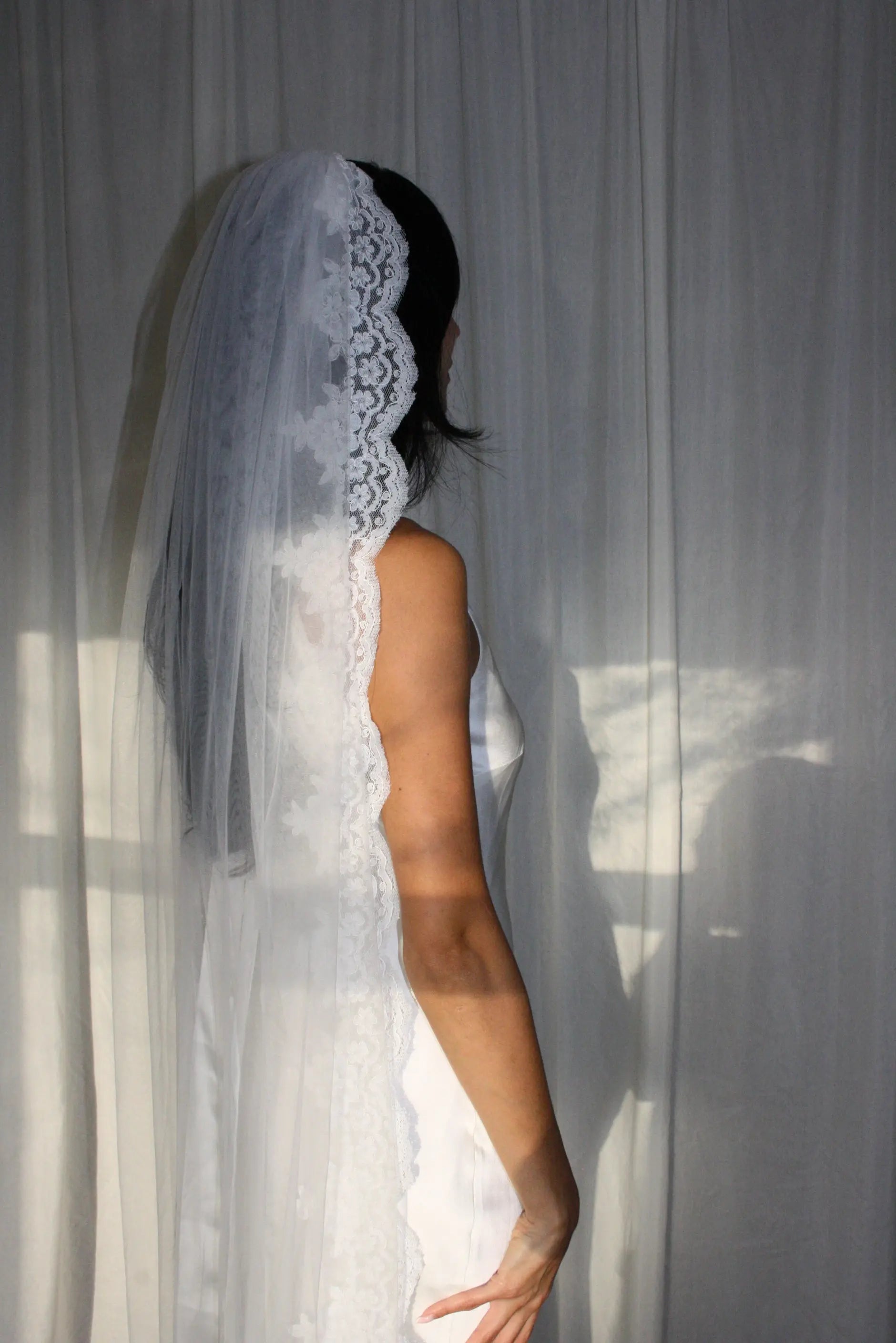 A woman shown from the back wearing a white bridal gown and a long sheer veil with a floral lace border, standing against a white curtain with soft sunlight and shadows.