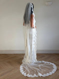 Load image into Gallery viewer, A woman wearing a white wedding dress and long sheer lace veil stands in profile in a room with a light wall and wood floor. The dress has a fitted bodice, cap sleeves, and a dramatic circular lace-trimmed train.
