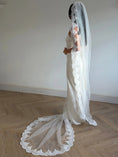 Load image into Gallery viewer, A woman in a white wedding dress and long sheer veil with lace trim stands in profile in a room with light wood herringbone flooring and a plain off-white wall.

