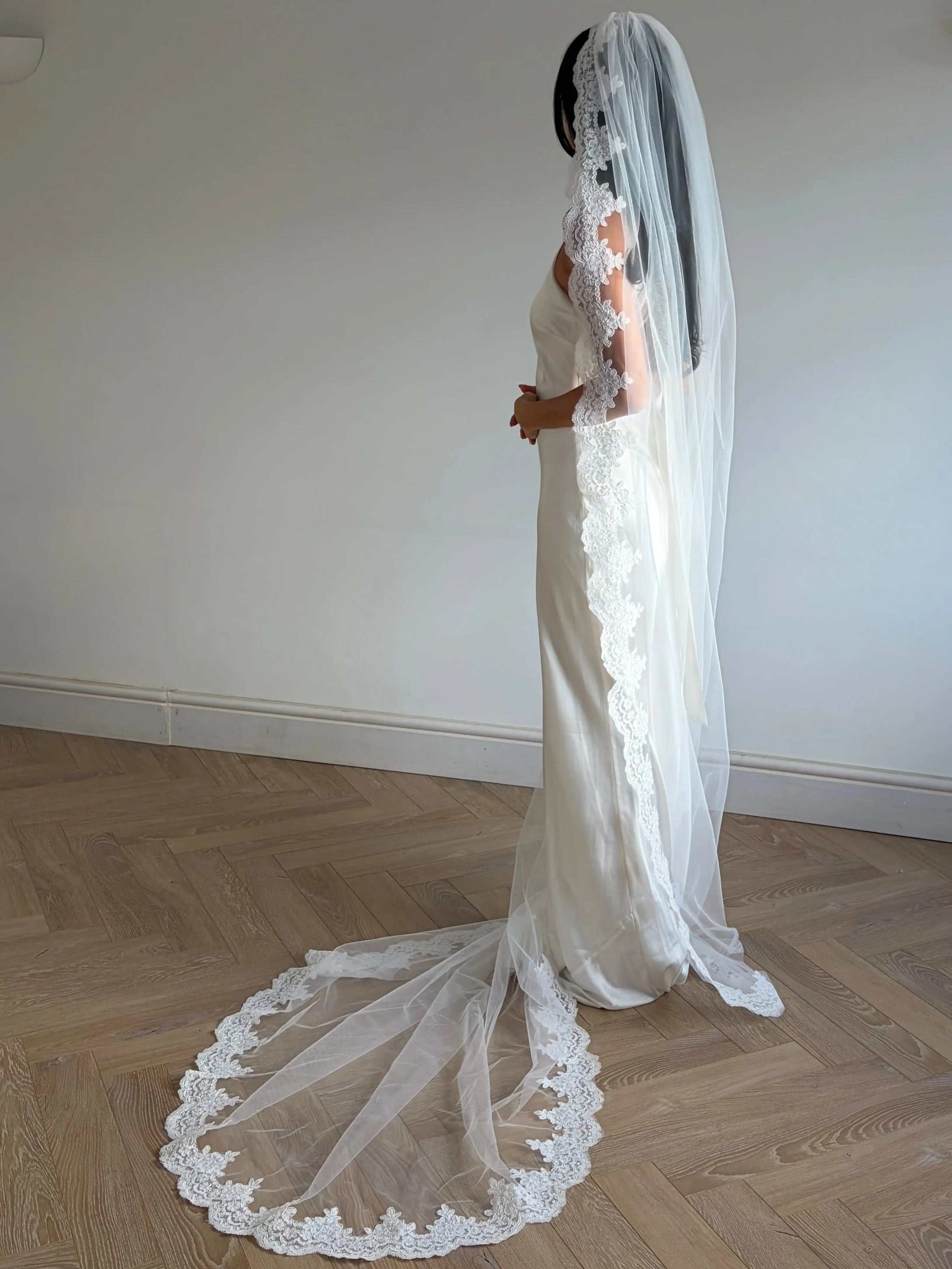 A woman in a white wedding dress and long sheer veil with lace trim stands in profile in a room with light wood herringbone flooring and a plain off-white wall.