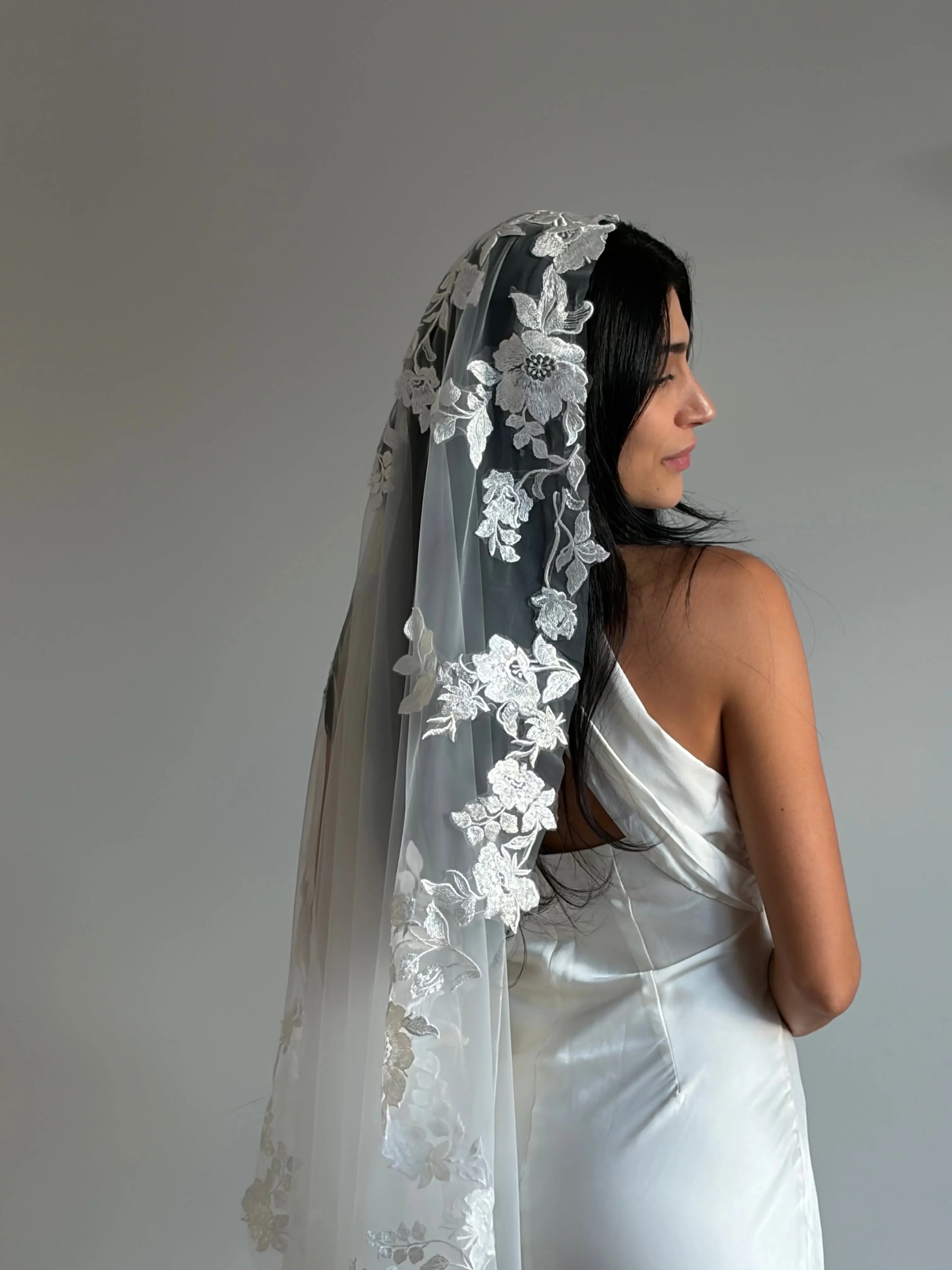 A woman in a white bridal gown shown from the back and side, wearing a long sheer white floral lace veil against a plain light gray studio background.