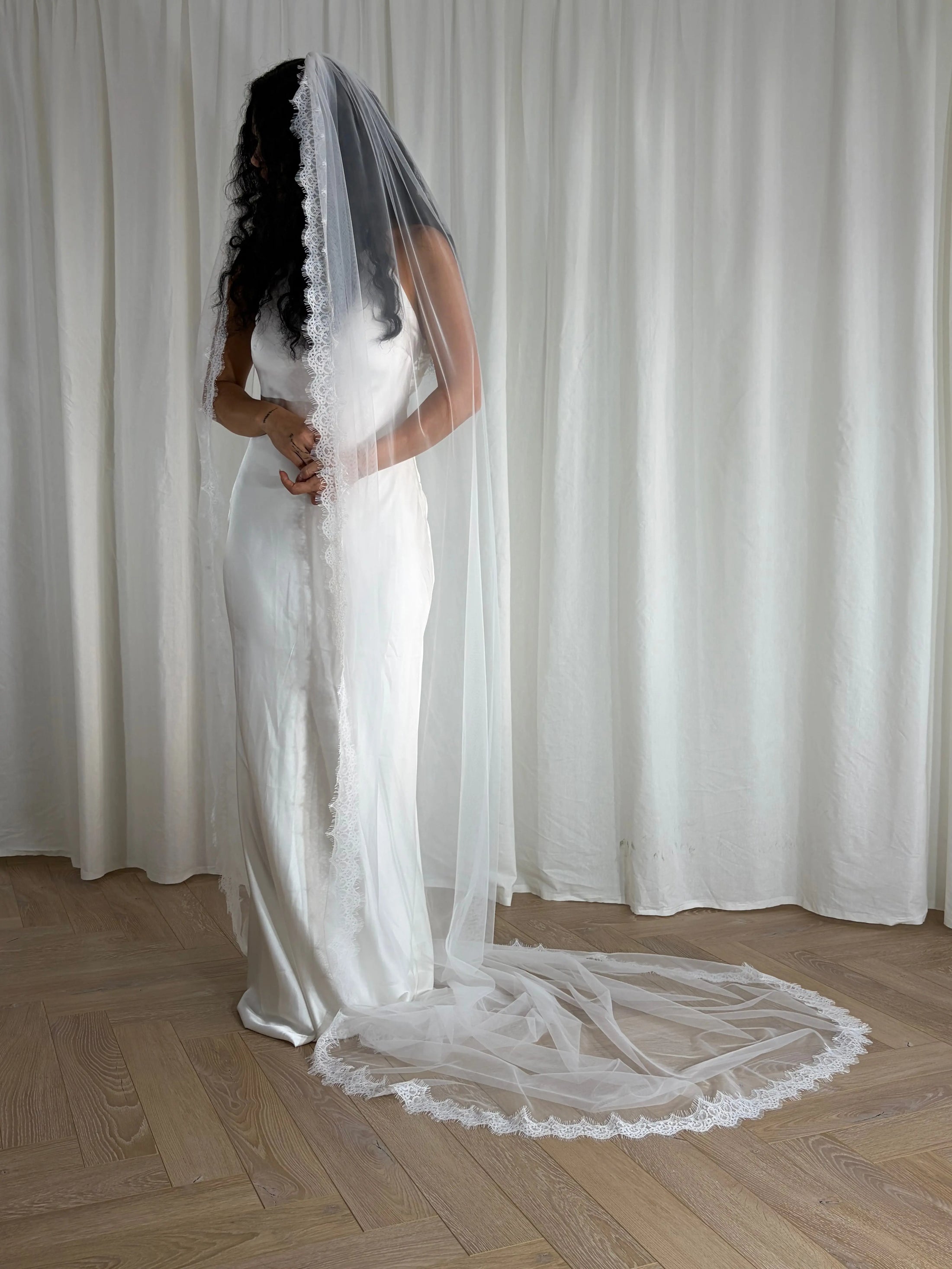 Person wearing a white wedding dress with a long veil in front of white curtains.